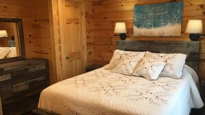Ein Schlafzimmer einer Holzhütte mit einem Bett, zwei Nachttischlampen, einer Kommode und einer Tür.