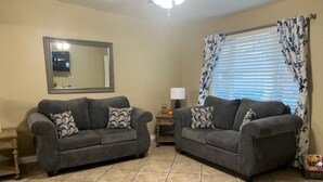 A living room with two grey sofas, patterned cushions, a small table with a lamp, a mirror on the wall, and a window with blinds and curtains. - Nice 3 bedroom's house owner occ and 2 bathroom with fast WI-Fi,  Full Kitchen (Las Vegas)