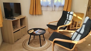 Een woonkamer met een televisie, twee stoelen, een klein tafeltje met een tijdschrift en een houten kast.