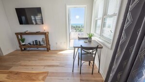 A room with a wooden floor, a table with chairs, a TV mounted on the wall, and a window with blinds. - Updated Oceanview Modern Decor, Private Balcony! (North Myrtle Beach)
