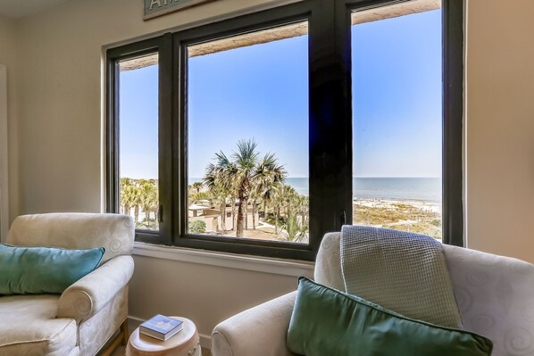 A room with two white armchairs, a small round table, and a view of the beach through large windows.