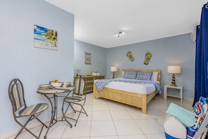 Ein Schlafzimmer mit einem Bett, zwei Nachttischen mit Lampen, einem Sessel, einem kleinen Tisch mit Obstschale und einer Wand mit einem Poster zum Thema Strand sowie einem Schild mit der Aufschrift 'Hawaii BBQ'.
