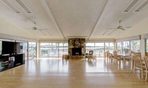 Relax in this spacious living room with a high ceiling and large windows offering a view of the surrounding area. Features a stone fireplace, flat-screen TV, and dining area with a table and chairs.