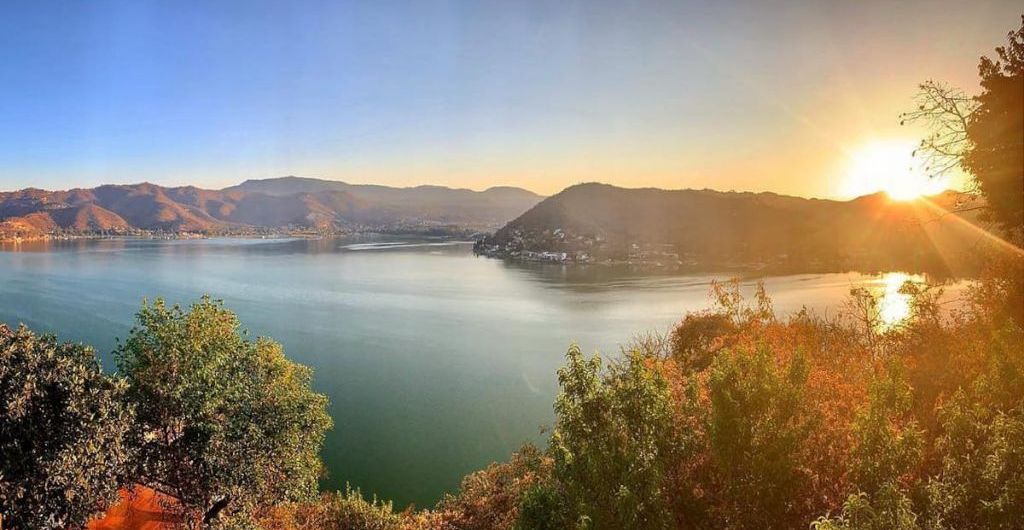 A panoramic view of a lake with mountains in the background and a sunrise or sunset on the horizon.