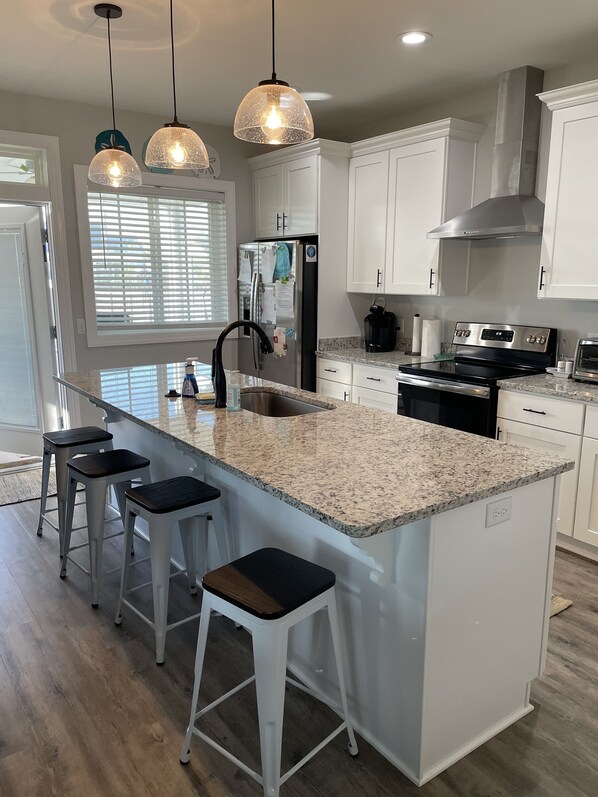 A modern kitchen with white cabinetry, a large island with bar stools, stainless steel appliances, and pendant lighting. - Super Cute Beach House— our slice of heaven! (Port St. Joe, Florida)