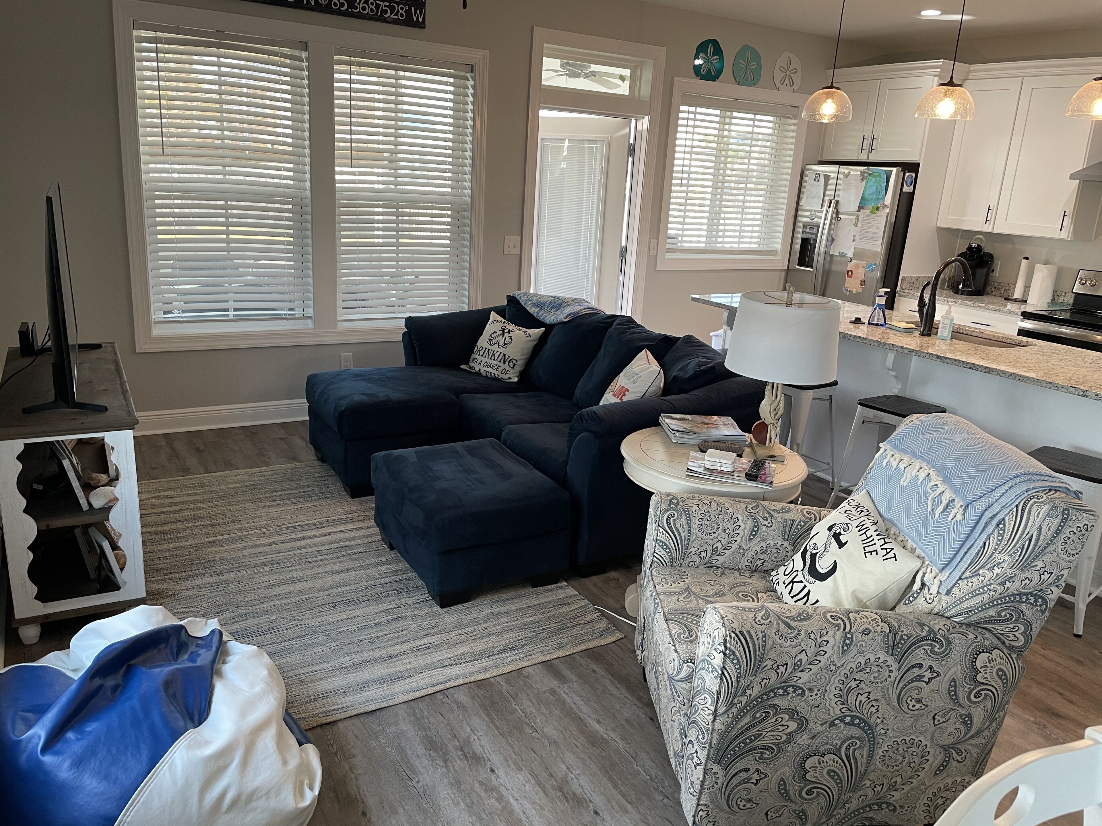A living room with a sectional sofa, a patterned armchair, a small round table, and a kitchen area with cabinets and appliances.
