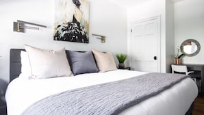 A modern bedroom with a large bed, a painting on the wall, a desk with a chair, and a door. - M1 at Saint Philip Square, One Block to King, Cozy Bright Stay (Charleston)