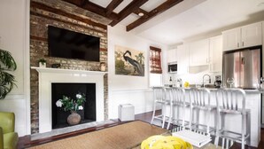 A living room with a fireplace, a dining area with white chairs, and a kitchen with white cabinets and a stainless steel refrigerator. - M1 at Saint Philip Square, One Block to King, Cozy Bright Stay (Charleston)