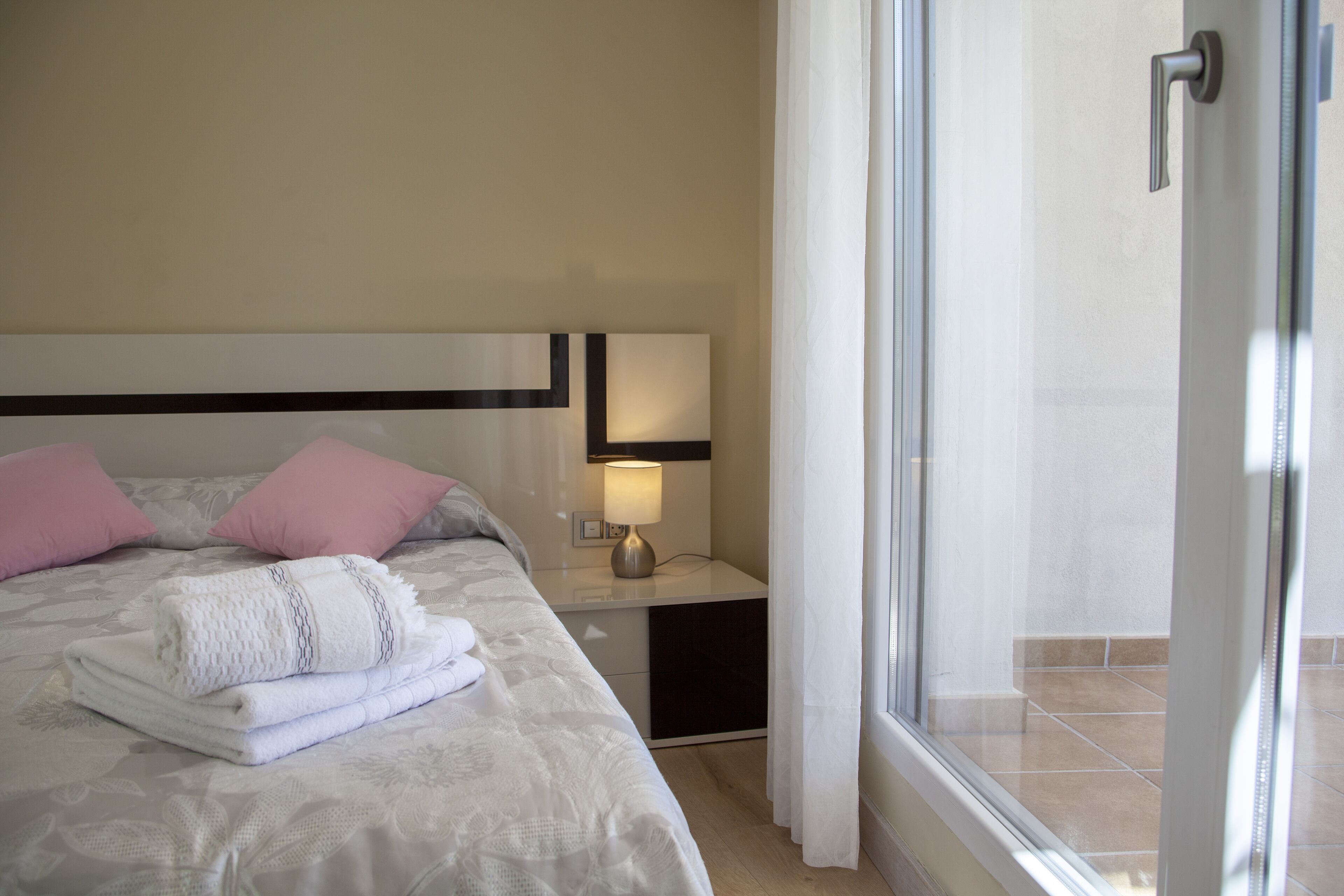 A neatly made bed with pink pillows, a bedside lamp, and a view of a tiled balcony through a glass door.