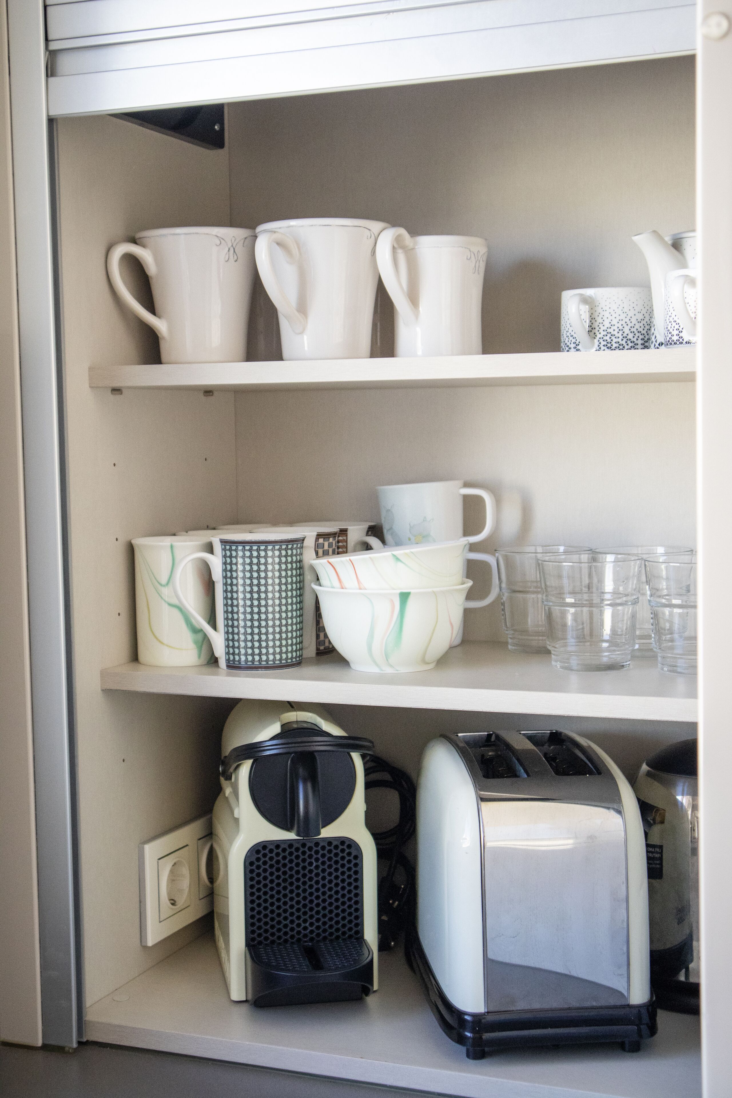 Una cocina con estantes blancos que sostienen diversos utensilios de cocina, incluyendo tazas, vasos y un tostador.