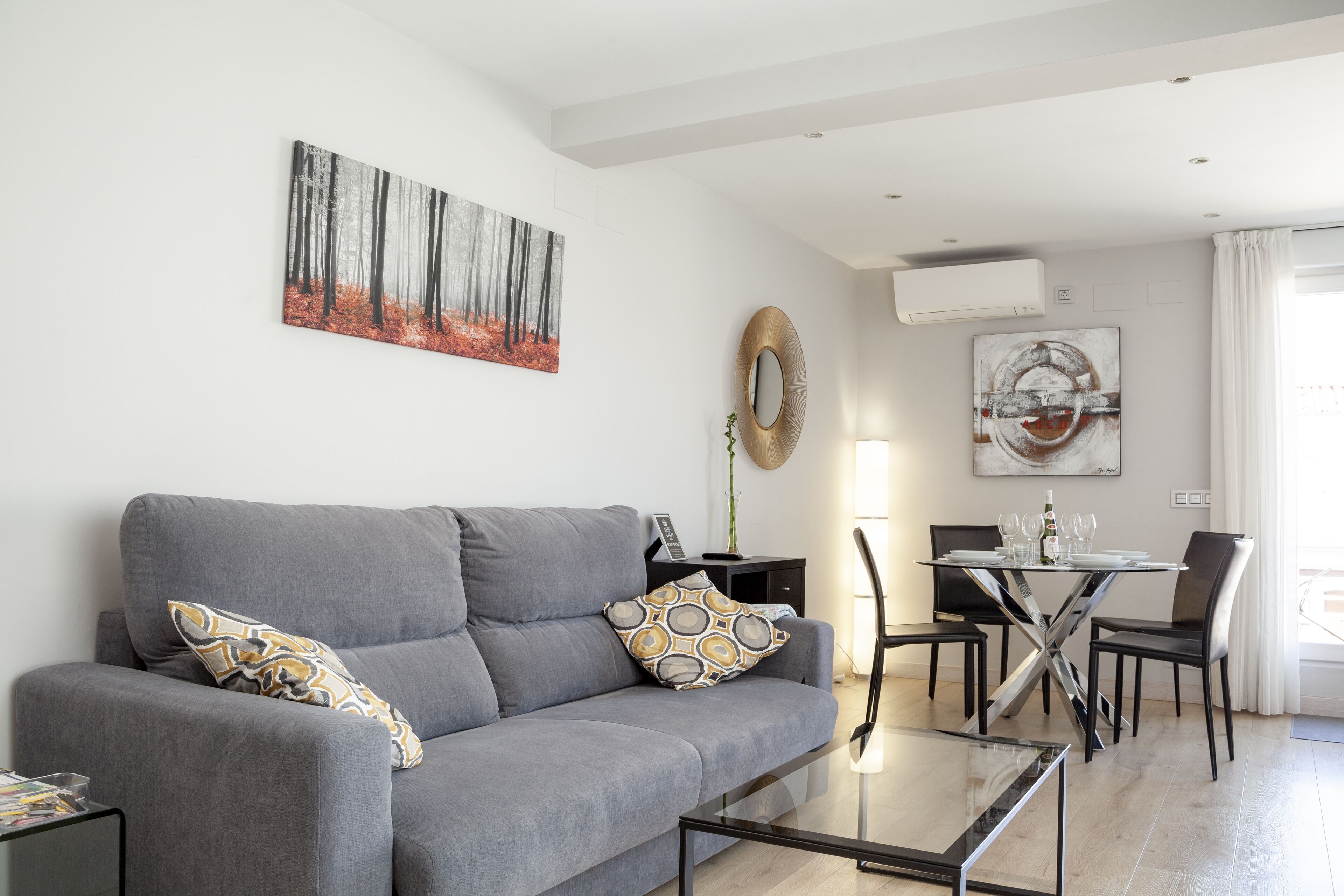 Una sala moderna con un sofá gris, cojines estampados, una mesa de centro de vidrio y un comedor con una mesa blanca y sillas negras.