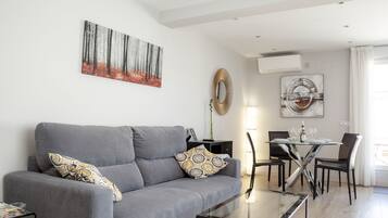 Una sala moderna con un sofá gris, cojines estampados, una mesa de centro de vidrio y un comedor con una mesa blanca y sillas negras.