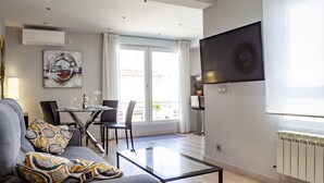 A modern living room with a sofa, a glass coffee table, a dining area with a table and chairs, a TV mounted on the wall, and a large window with curtains.