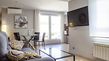 Ett modernt vardagsrum med en soffa, ett soffbord i glas, en matplats med bord och stolar, en tv som är monterad på väggen och ett stort fönster med gardiner.