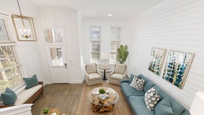Un salón amplio con un sofá modular, una mesita redonda y dos sillones. Se observa una puerta con ventana, una pequeña mesa con lámpara y una obra de arte colgada en la pared.
