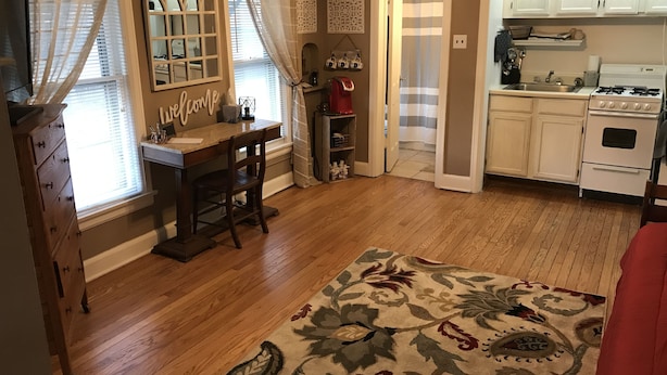 Una habitación con piso de madera, un escritorio con silla, una ventana con cortinas, una cocina con gabinetes blancos y una estufa, y un tapete con estampado floral.