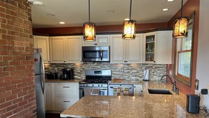 Uma cozinha com armários brancos, bancada de granito, fogão de aço inoxidável e micro-ondas. Duas luminárias pendentes iluminam a ilha.