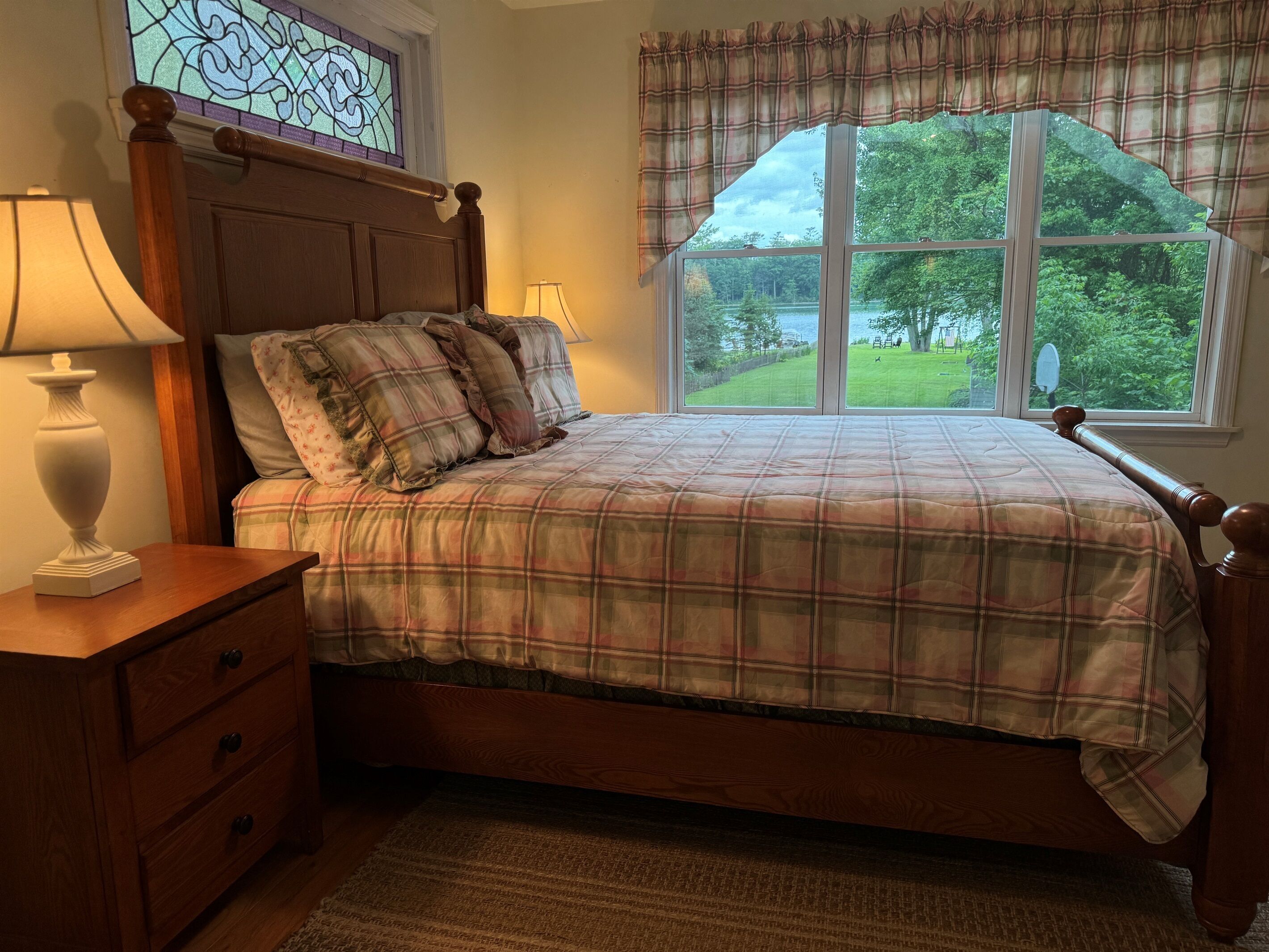 Un dormitorio con una cama de madera, ropa de cama escocesa, una mesita de noche con lámpara, un aparador y una ventana con vistas a la vegetación.