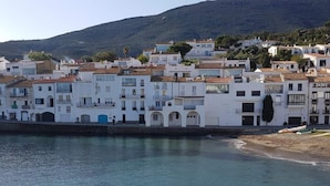 A coastal town with white buildings, a clear blue sea, and a hilly backdrop.