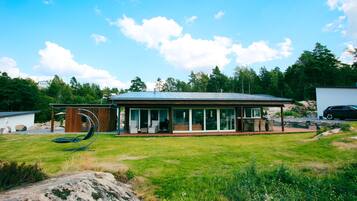 Une maison moderne dotée d’une grande façade vitrée, d’un patio avec mobilier d’extérieur et d’une pelouse verdoyante tout autour.