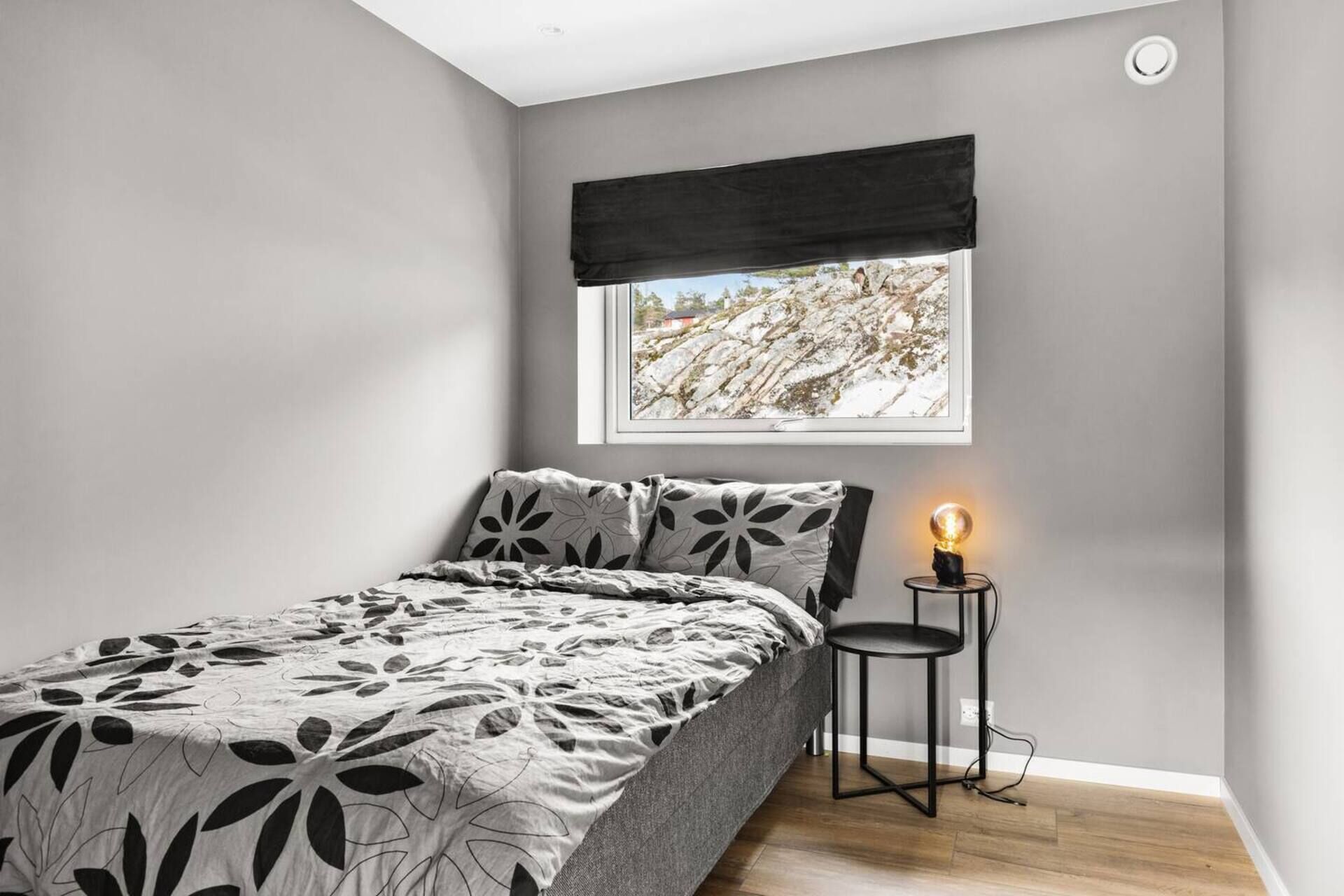 A bedroom with a bed, a nightstand with a lamp, a window with a view of a rocky landscape, and a black curtain.