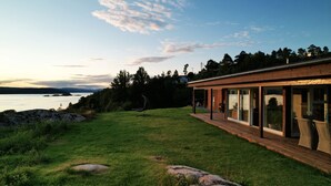 A modern house with a deck overlooking a lake, featuring a glass-enclosed patio and a hanging chair. - Cottage - Sleeps 14 With Fjord Views (Frogn)