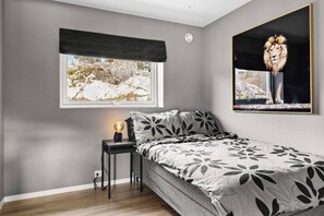 Une chambre à coucher avec un lit, une table de chevet, une lampe, une fenêtre donnant sur des arbres et un tableau encadré représentant un lion.