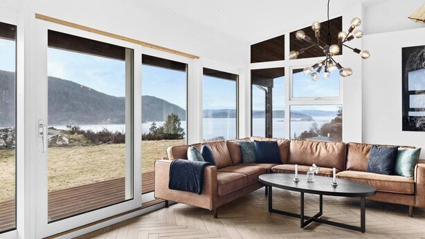 A modern living room with a leather sectional sofa, a round coffee table, and large floor-to-ceiling windows offering a view of a lake and hills. - Cottage - Sleeps 14 With Fjord Views (Frogn)
