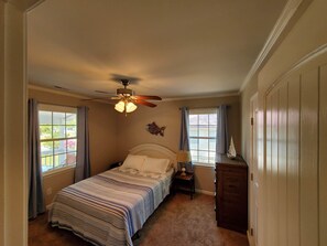 Um quarto com cama, mesa de cabeceira, luminária, ventoinha no teto e janela com cortinas.