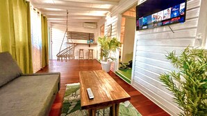 A cozy living room with a sofa, a wooden table, a hanging chair, a potted plant, and a TV mounted on the wall displaying a streaming service. - Jungle Cottage Orient Bay: Charming and authentic (Saint Martin)