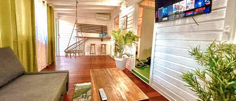 A cozy living room with a sofa, a wooden table, a hanging chair, a potted plant, and a TV mounted on the wall displaying a streaming service.