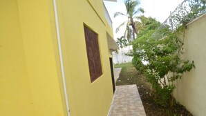 A narrow pathway leading to a yellow building with a small window and a white wall with a tree and a fence in the background. - Residencial Lakis 1 (Boca Chica)
