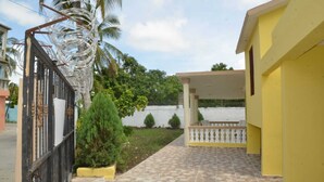 A gated residential area with a paved walkway, a covered porch, and a small garden with a palm tree and shrubs. - Residencial Lakis 1 (Boca Chica)