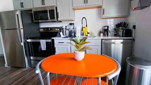 Una cucina compatta con elettrodomestici in acciaio inossidabile, un piccolo tavolo da pranzo con sedie arancioni e pensili bianchi.
