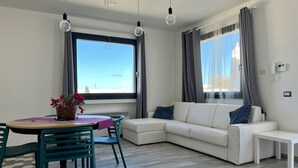 Ein modernes Wohnzimmer mit einem weißen Sofa, einem Esstisch mit Stühlen und einem großen Fenster mit Vorhängen.