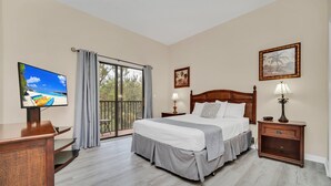 Une chambre d’hôtel avec un grand lit, une télévision, un balcon avec des rideaux et une table de chevet en bois avec une lampe.