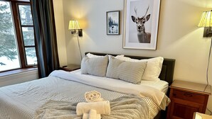 Una cama bien hecha con ropa de cama a rayas, un edredón mullido y una toalla con un logotipo. Hay dos lámparas de noche, un cuadro enmarcado de un ciervo y una ventana con cortinas.