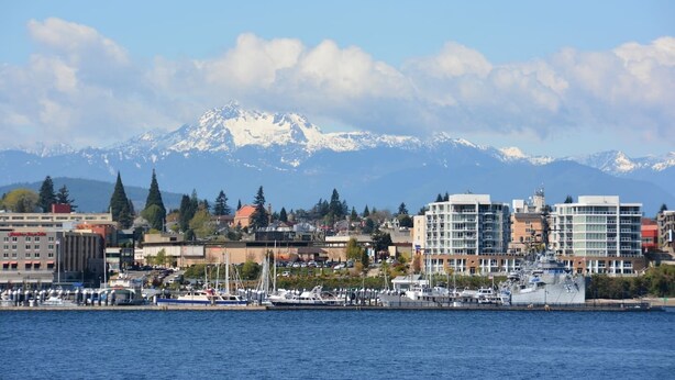 A coastal cityscape with modern buildings, a marina with boats, and snow-capped mountains in the background.
