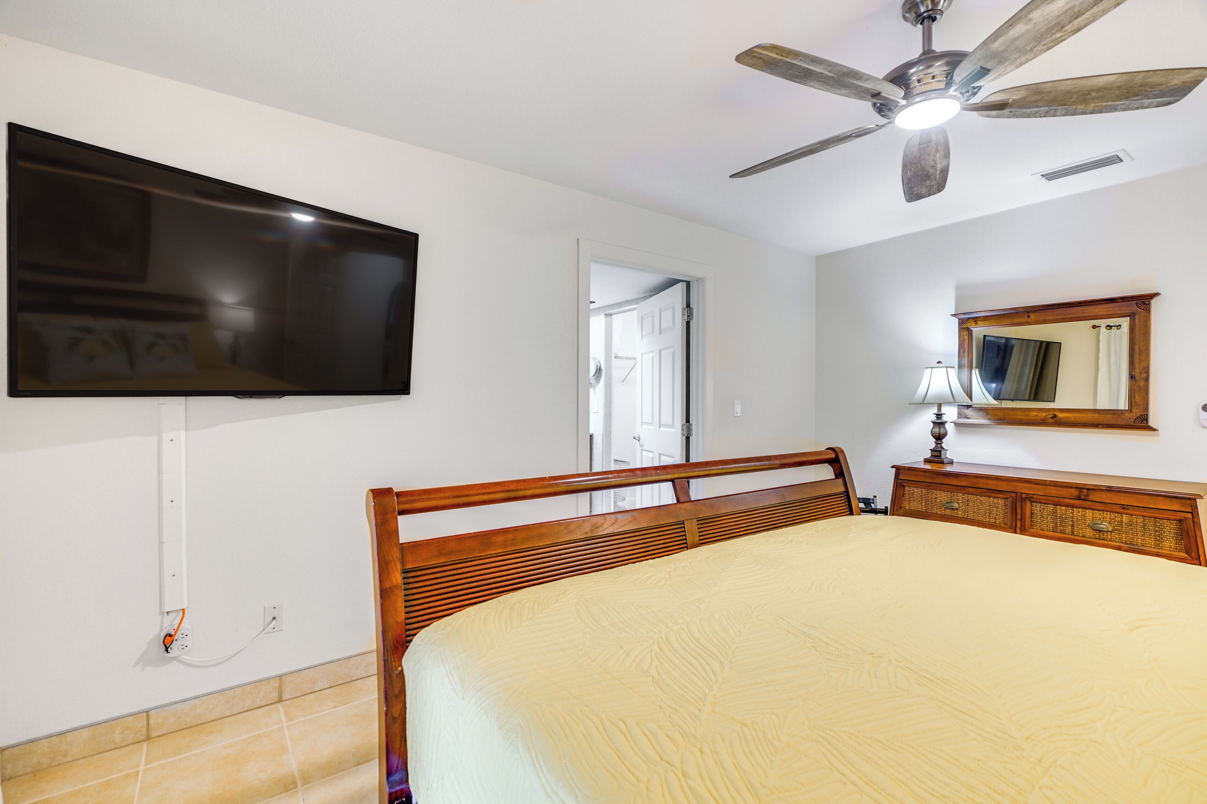 A bedroom with a bed, a television mounted on the wall, a ceiling fan, and a wooden dresser.