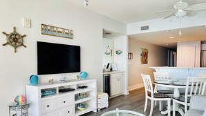 Una sala de estar moderna con televisor de pantalla plana, muebles blancos, una mesa de centro de vidrio y un comedor con sillas blancas.