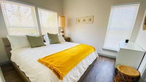 Une chambre à coucher avec un grand lit, du linge de lit blanc, un plaid jaune et un petit tabouret.