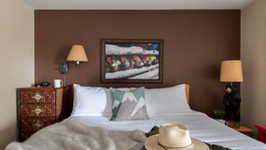 A neatly made bed with a white comforter, a beige hat, and a fur throw. There is a wooden nightstand with a clock and a lamp, and a wall-mounted lamp. A painting of a snowy landscape hangs above the bed. - Outpost: Pitchfork 432 - Close to Snow King Mtn! (Jackson)