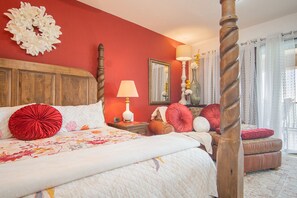 A bedroom with a four-poster bed, a wooden headboard, a red wall, a mirror, a bedside table with a lamp, and a window with curtains.