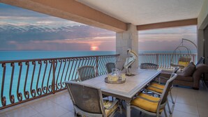 A balcony with a dining table, chairs, and a hanging chair overlooking the sea at sunset.