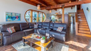 A spacious living area with a large L-shaped sectional sofa, a wooden coffee table with snacks, and a view of the kitchen through large windows. - Bayside Breeze | Waterfront Patio & Sunset Views (San Diego)