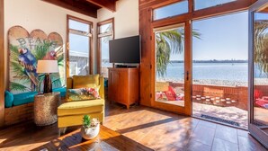 A living room with a large window overlooking a beach, a television, and a cozy seating area. - Bayside Breeze | Waterfront Patio & Sunset Views (San Diego)