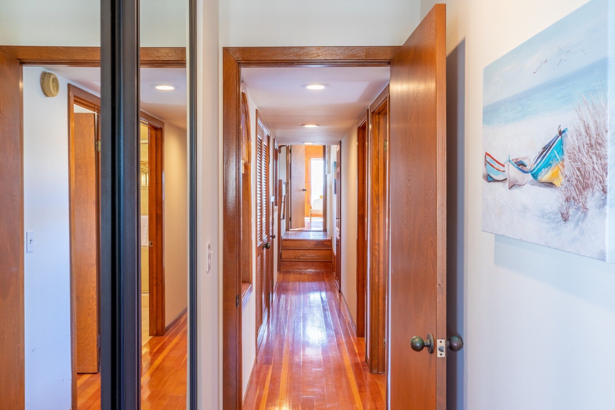 A hallway with wooden floors and doors, leading to a room with a painting of boats on the wall.