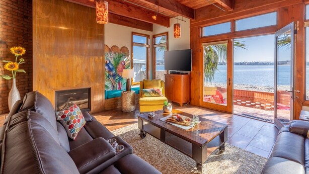 A spacious living room with a large sofa, a coffee table, and a TV. There is a fireplace, a painting, and a view of the beach through large windows. - Bayside Breeze | Waterfront Patio & Sunset Views (San Diego)