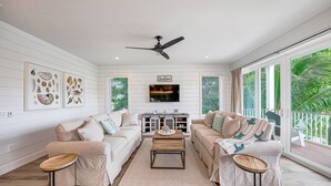 A spacious living room with two beige sofas, a wooden coffee table, and a ceiling fan. A flat-screen TV is mounted on the wall, and there is a large window with a view of greenery outside.