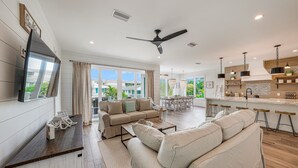 A spacious living room with a large sofa, a coffee table, and a TV mounted on the wall. There is a dining area with a table and chairs, and a kitchen area with a bar and stools.